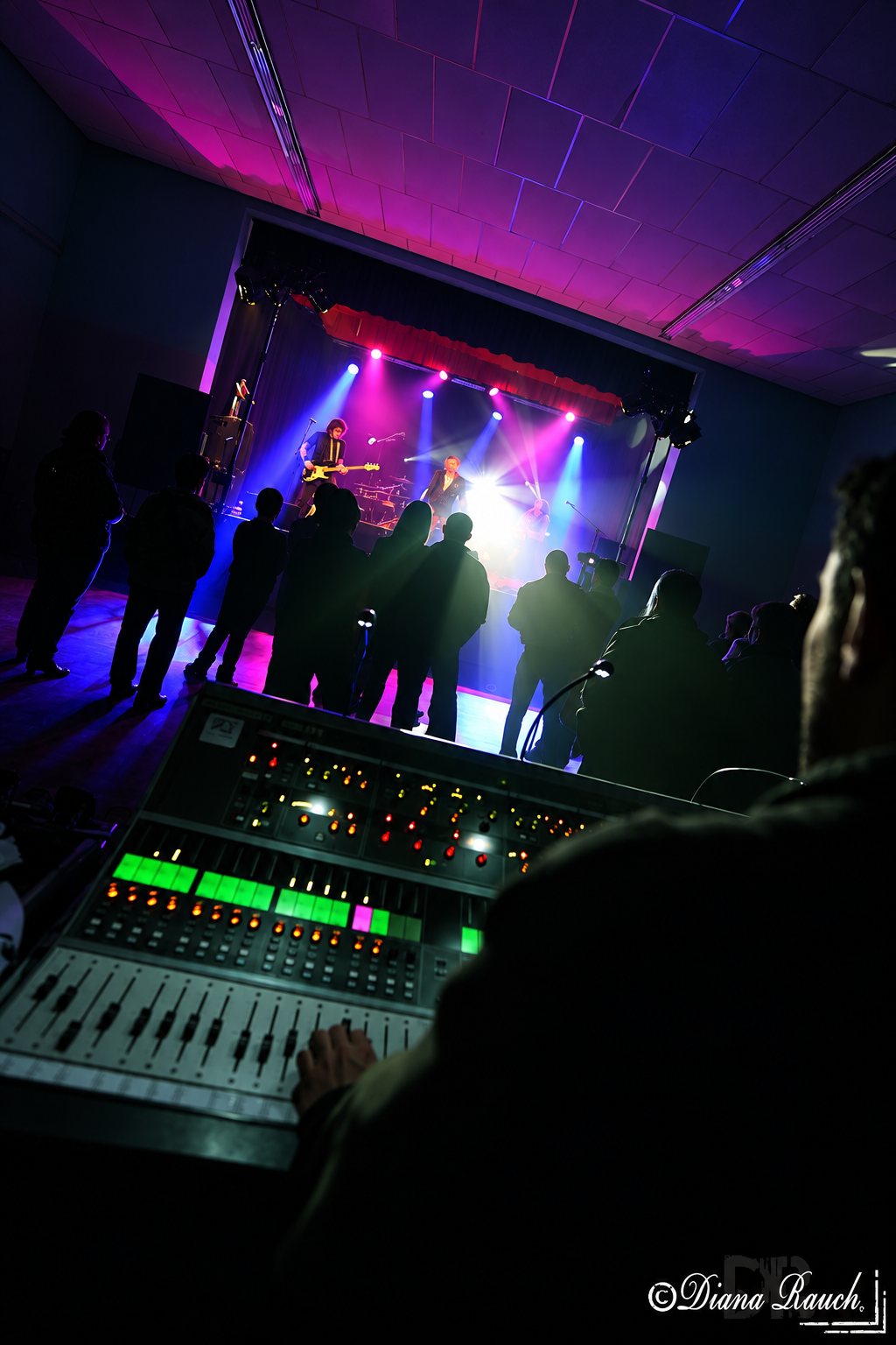 Un groupe joue sur scène sous des projecteurs violets et bleus, vu depuis la console de mixage, devant un public en silhouette.