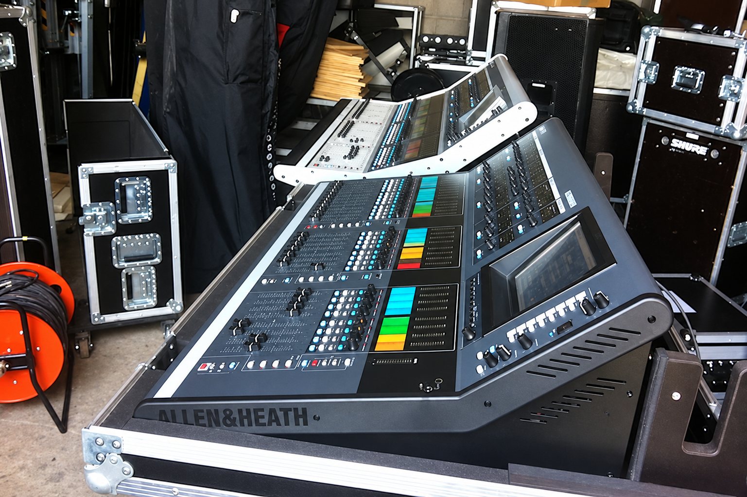Une grande console de mixage numérique Allen & Heath posée dans son flight case, entourée de caisses de transport et d'un enrouleur de câble orange dans un entrepôt.