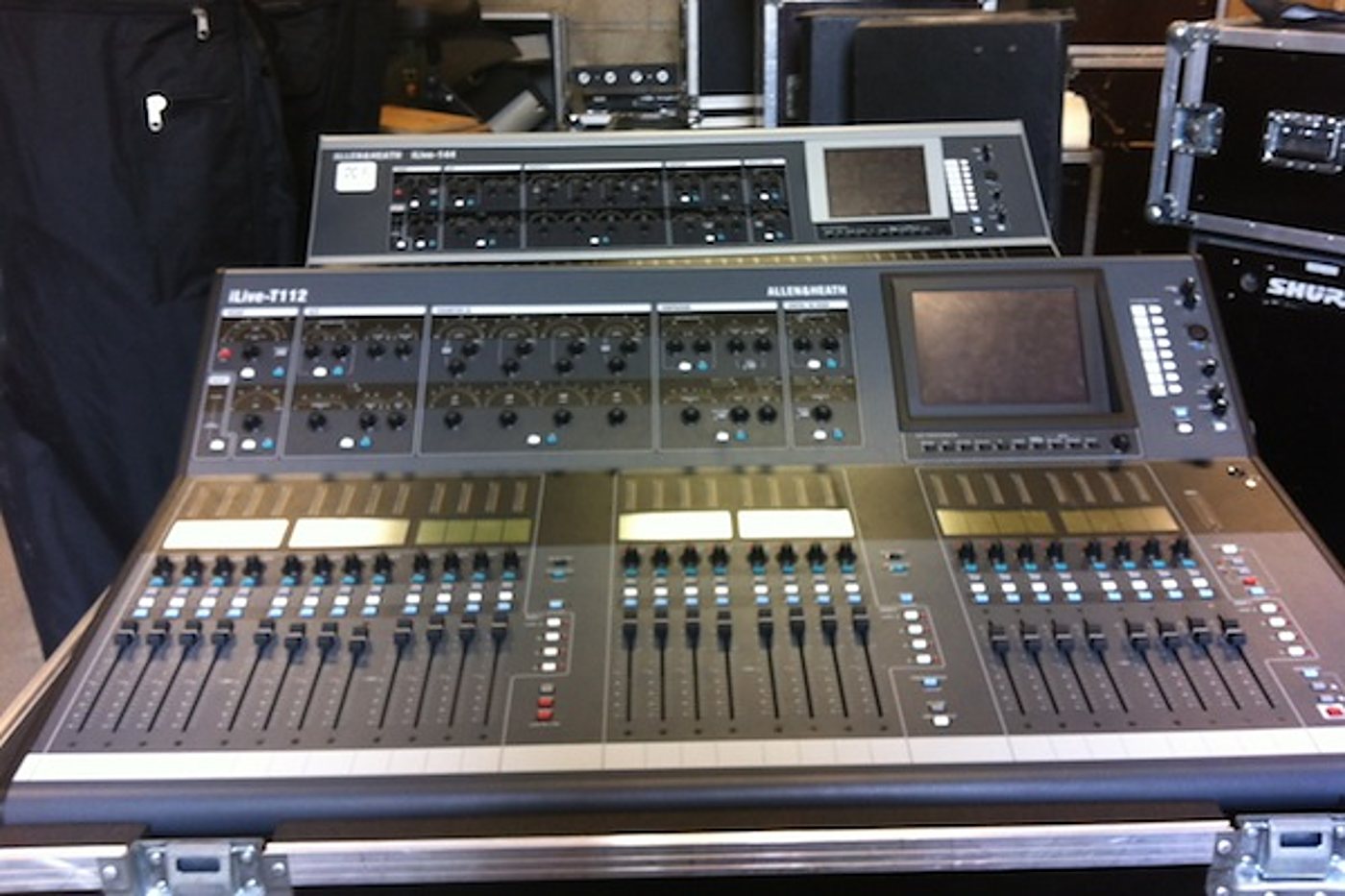Une console de mixage numérique Allen & Heath iLive-T112 posée sur son flight case, avec une seconde console derrière elle dans un local technique.