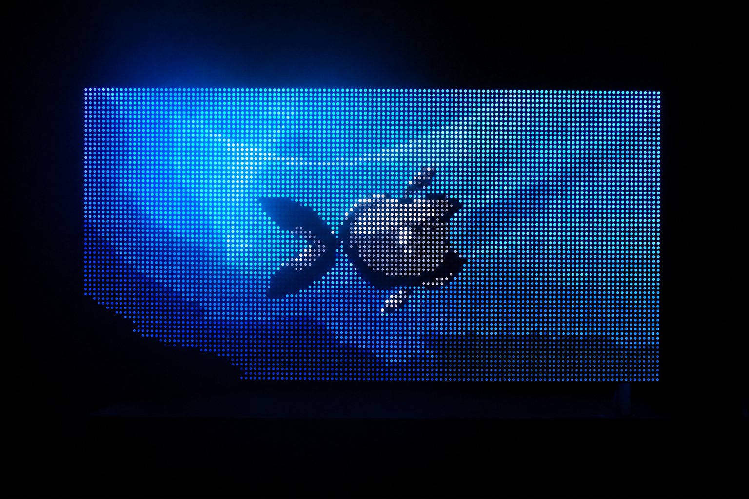 Un grand écran LED dans l'obscurité affiche un logo en forme de pomme stylisée sur un fond bleu dégradé, avec une trame de pixels bien visible.