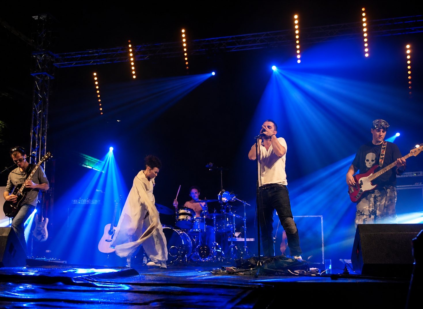 Un groupe de rock joue sur scène dans la pénombre, avec deux guitaristes, un chanteur à l'harmonica, un batteur et un danseur enveloppé de blanc sous des projecteurs bleus.