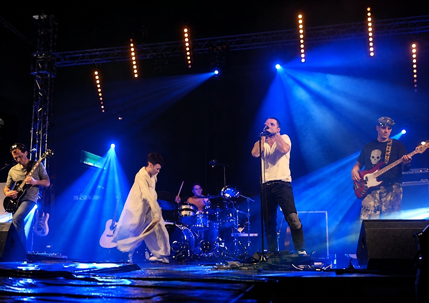 Cinq musiciens se produisent sur une scène éclairée de projecteurs bleus, avec deux guitaristes, un chanteur, un batteur et un musicien en tenue blanche.