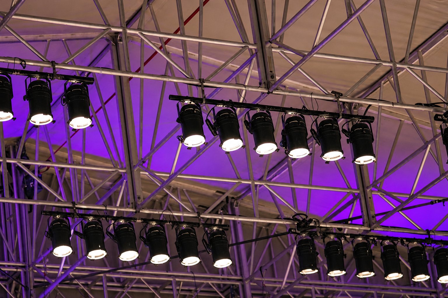 Plusieurs rangées de projecteurs allumés sont fixées à une structure métallique en treillis sous un plafond de chapiteau éclairé en violet.
