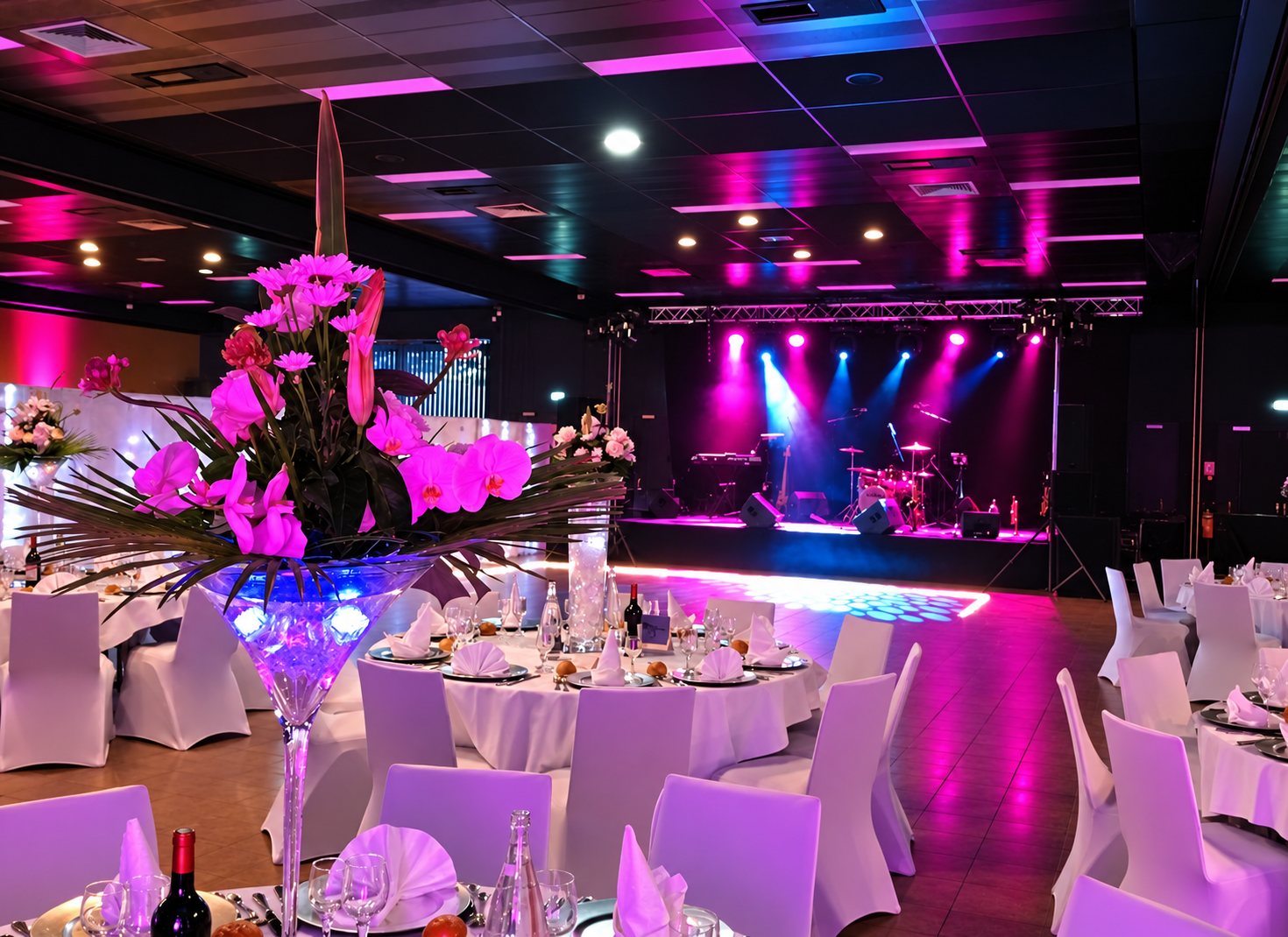 Une grande salle de réception aux tables rondes dressées, avec un haut vase fleuri au premier plan et une scène musicale éclairée de projecteurs roses et bleus au fond.