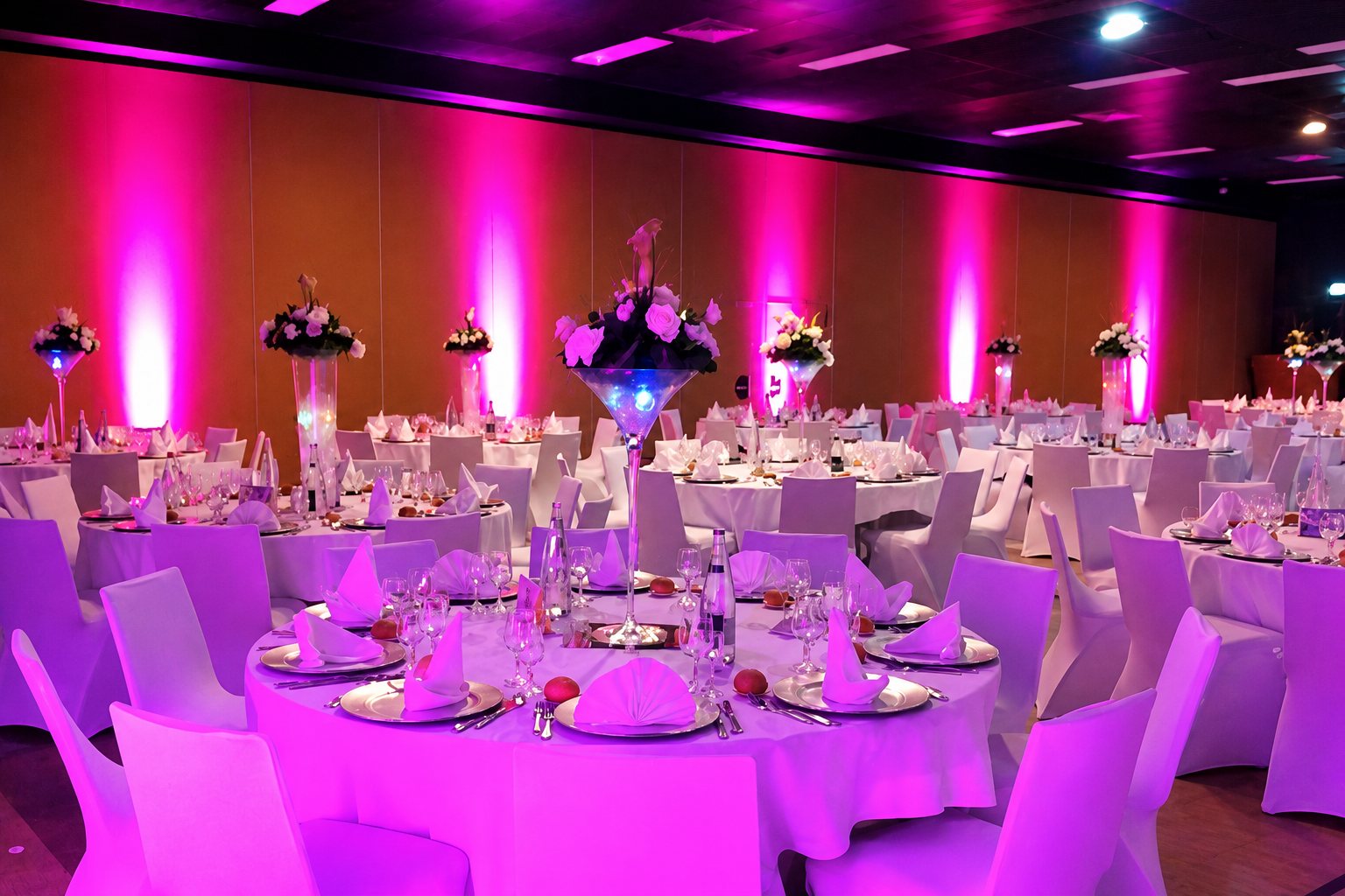 Grande salle de réception aux murs éclairés en rose et violet, avec des tables rondes nappées de blanc, des housses de chaises blanches et des centres de table floraux.