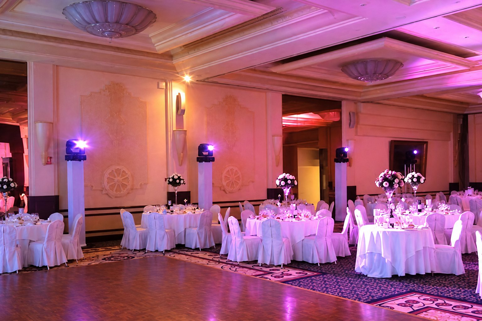 Grande salle de réception aux tables rondes nappées de blanc, chaises housssées et centres de table fleuris, éclairée de lumière rose et violette.