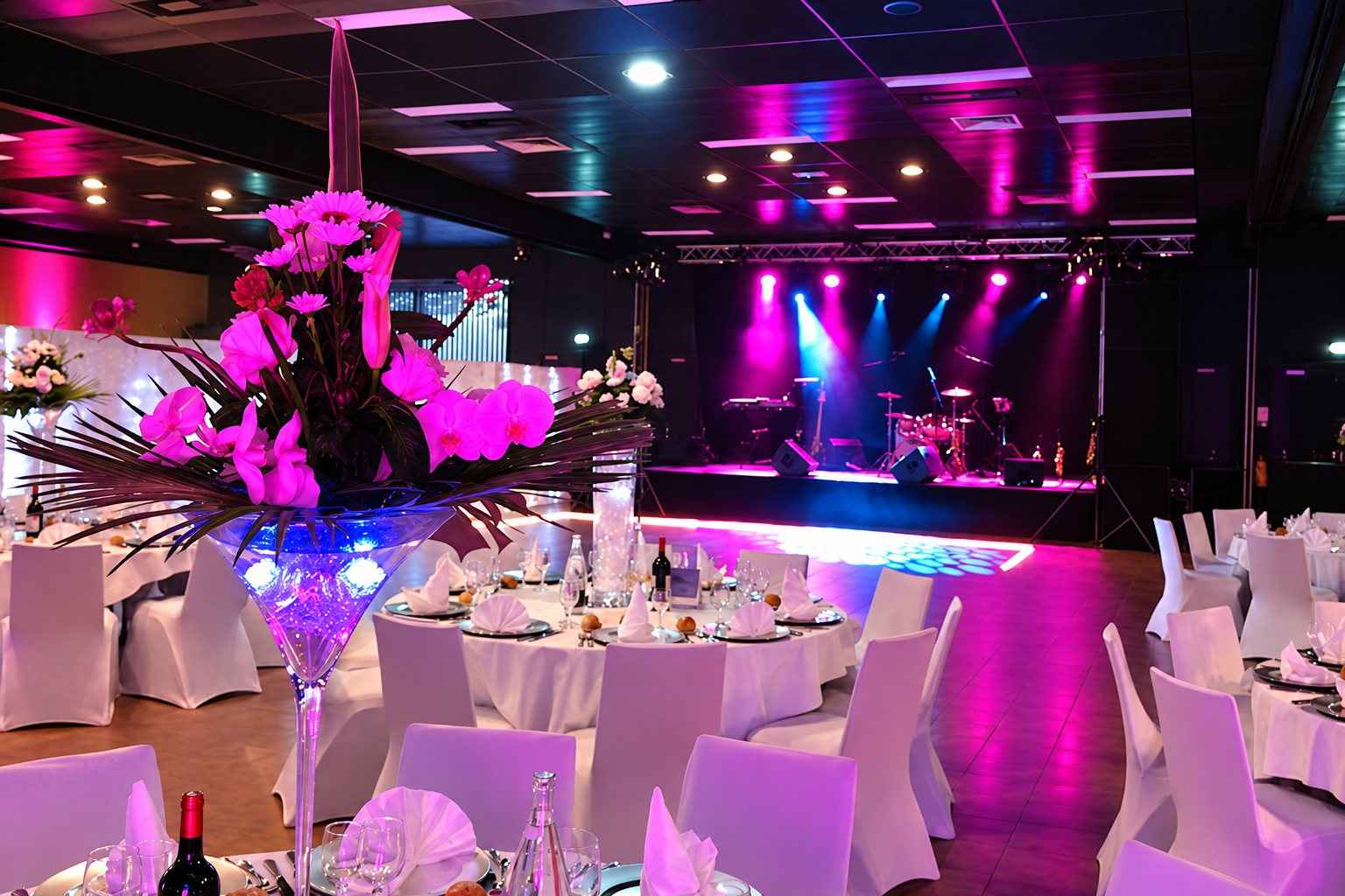 Grande salle de réception aux tables dressées avec housses blanches, un grand centre de table fleuri au premier plan et une scène illuminée de lumières roses et bleues au fond.