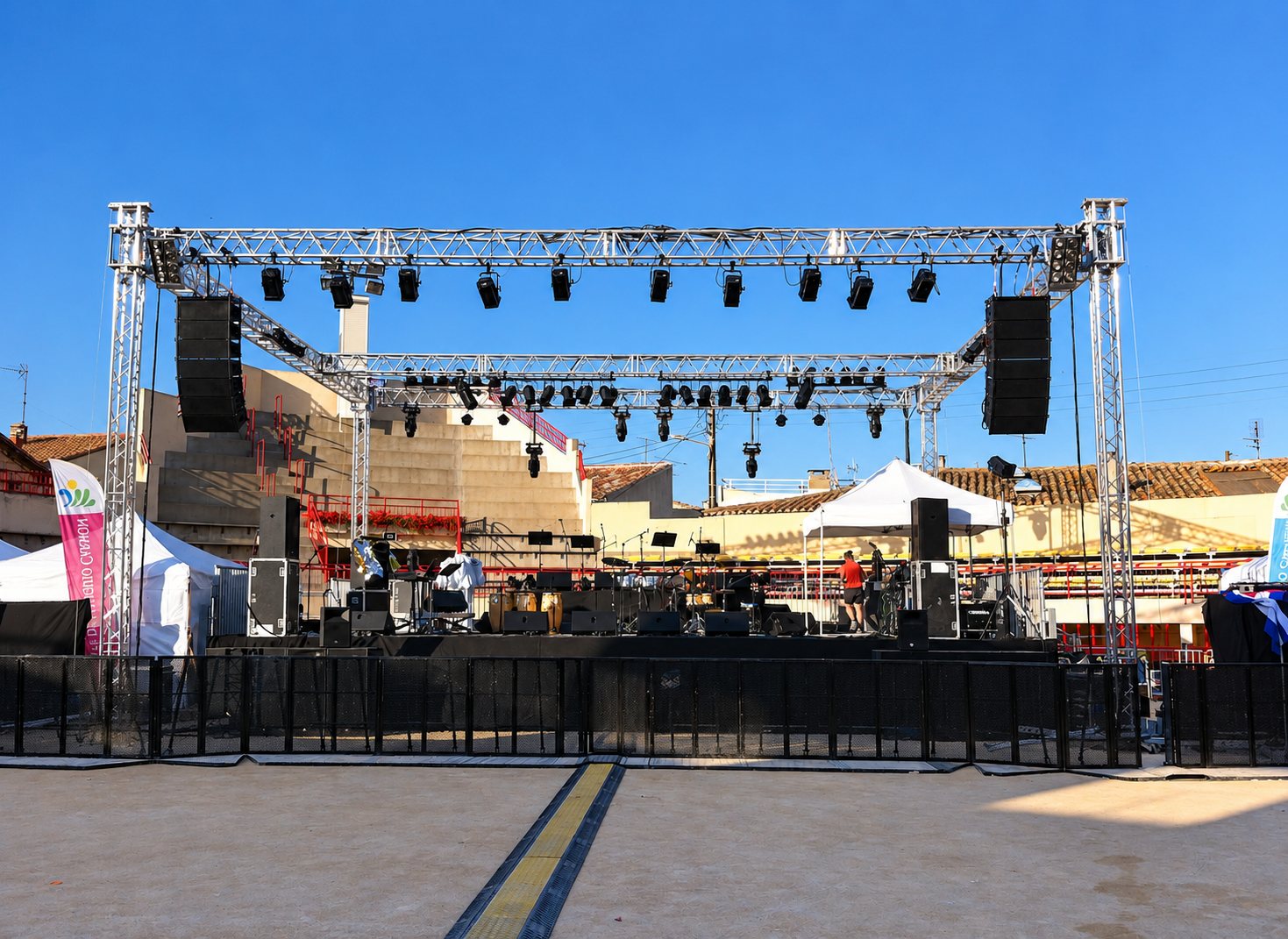 Une scène de concert en plein air est montée sous un ciel bleu, avec une structure en treillis métallique, des projecteurs, de grandes enceintes et des tentes blanches sur les côtés.