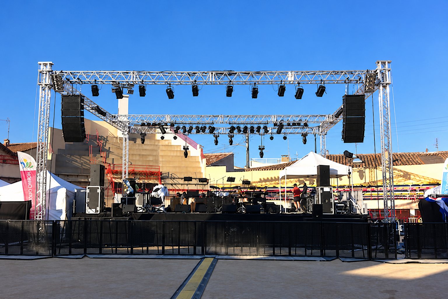 Une grande scène de concert en plein air équipée d'une structure en treillis métallique, de projecteurs et d'enceintes suspendues sous un ciel bleu.