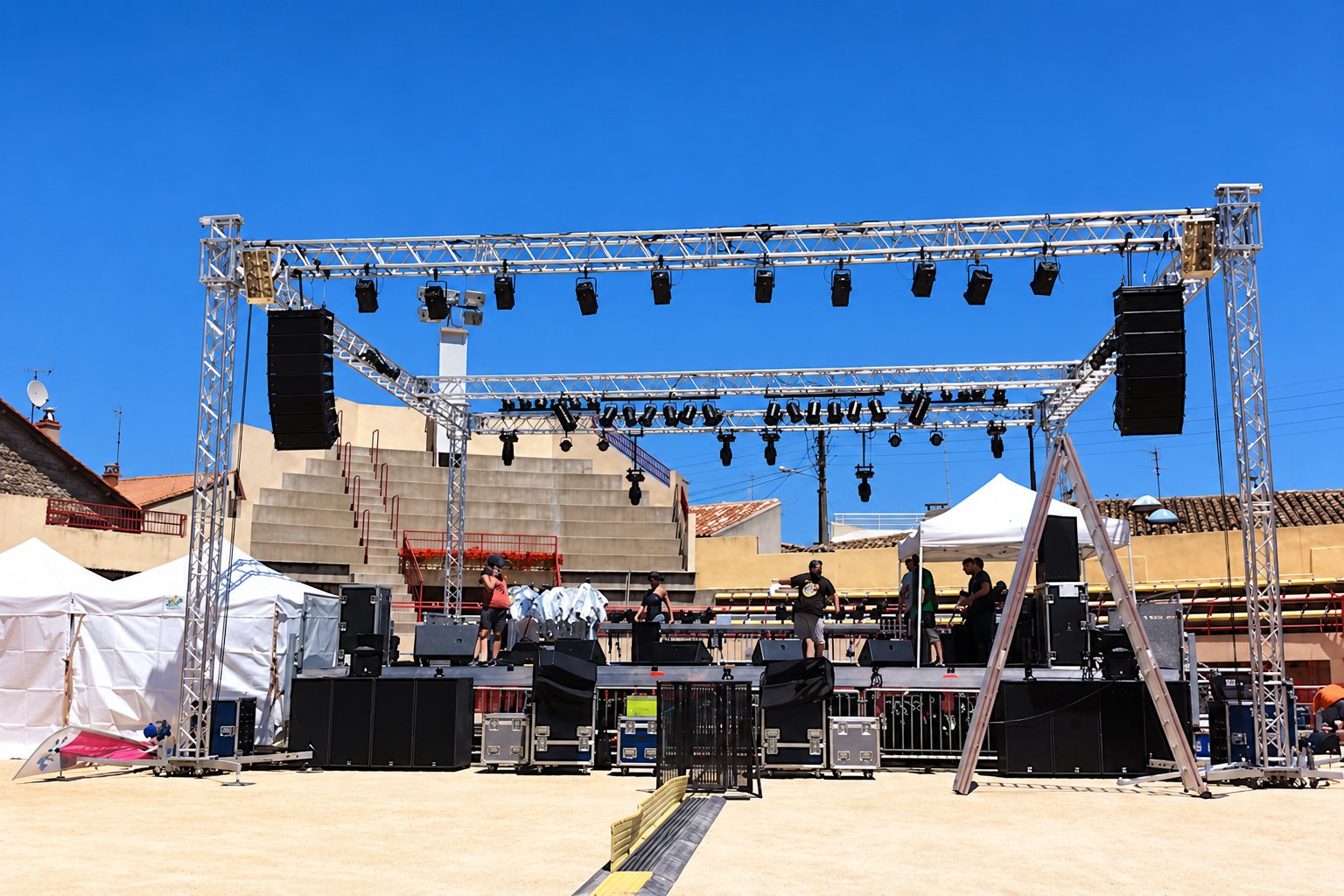 Une structure métallique en treillis supportant des projecteurs et de grandes enceintes est montée sur une scène en plein air, sous un ciel bleu, avec des techniciens au travail.