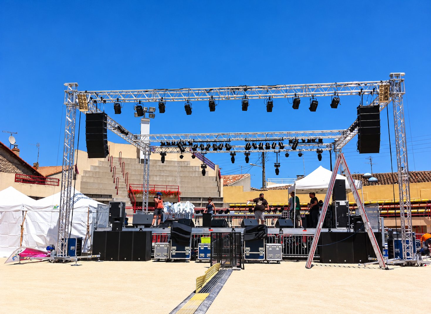 Des techniciens montent une scène de concert équipée de structures en treillis, de projecteurs et de grandes enceintes, devant des gradins en plein soleil.