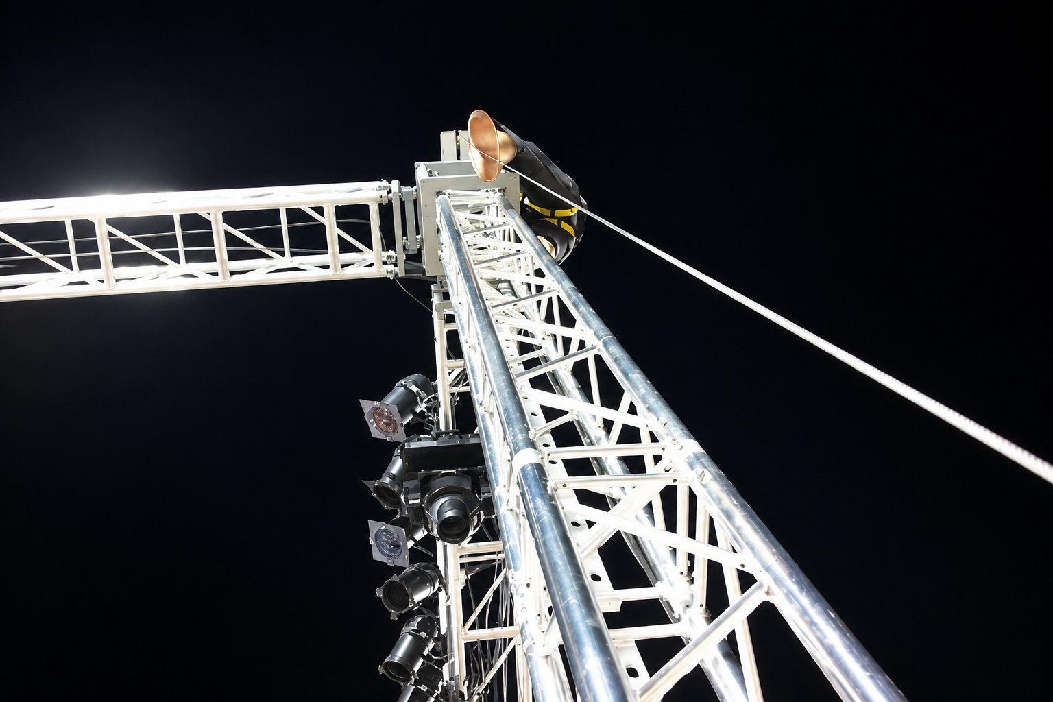 Vue en contre-plongée d'une haute structure en treillis métallique équipée de projecteurs, avec un technicien perché au sommet dans la nuit.